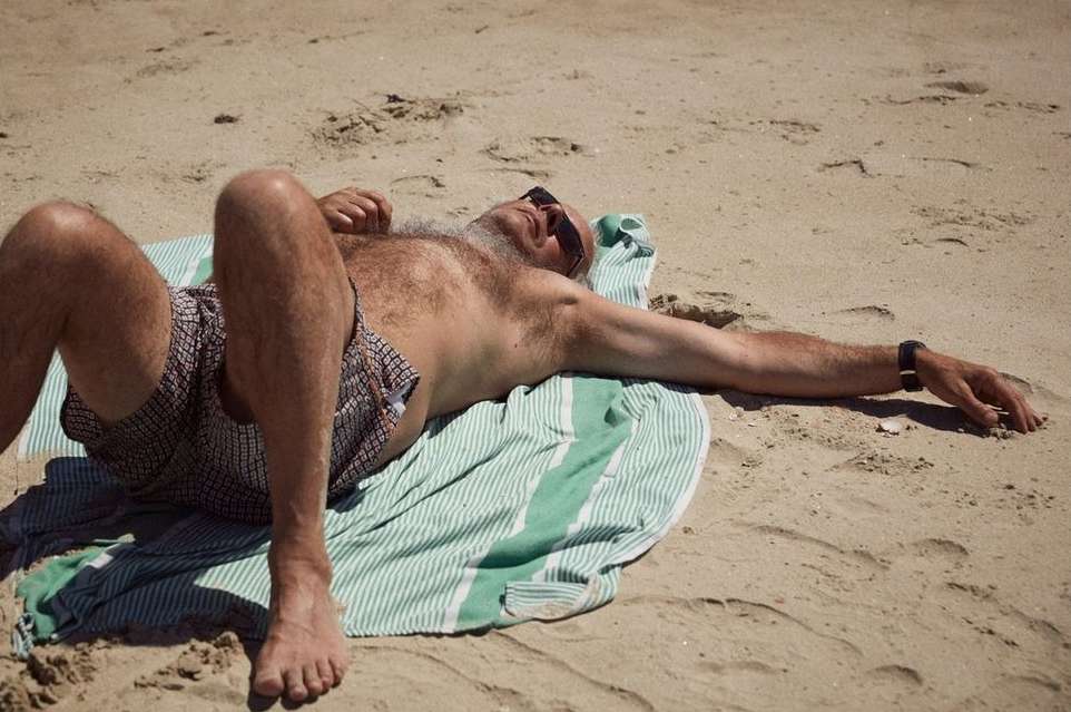 Mature man with grey beard and sunglasses laying on a striped towel at the beach, enjoying the sun on a bright summer day.