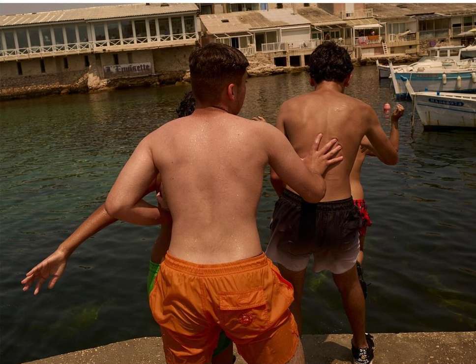 Four shirtless young men stand by the water's edge on a sunny day, with a building and boats visible in the background.
