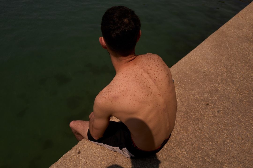 Rear view of a shirtless male sitting near dark water on a concrete structure, showcasing skin texture and natural outdoor light.