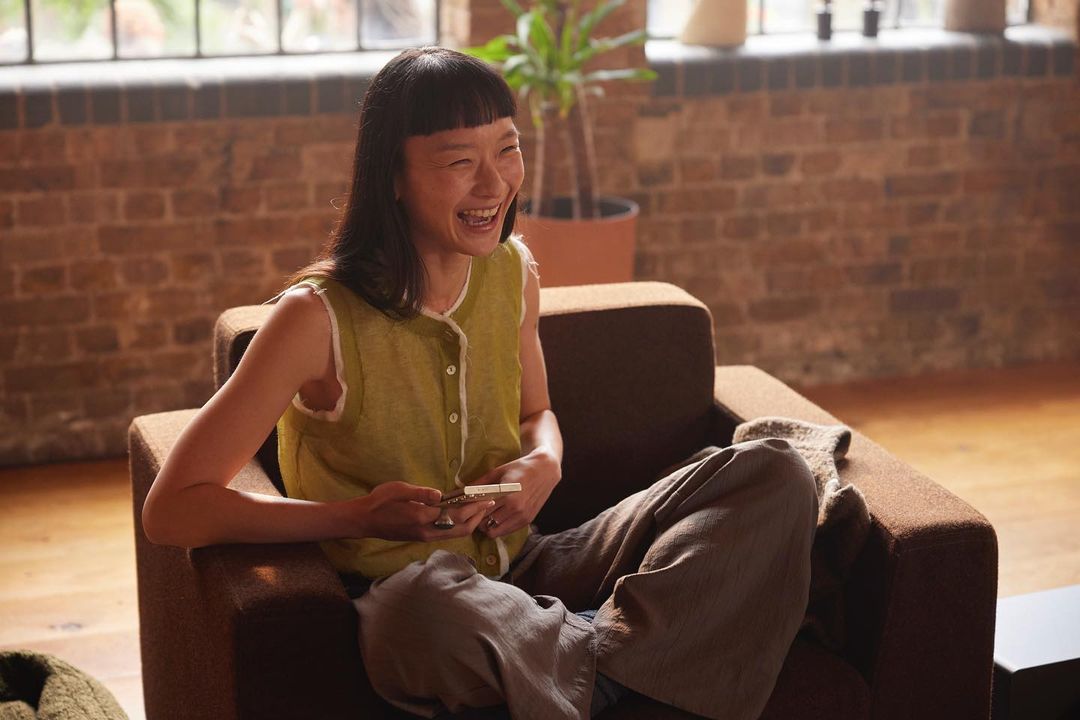 An Asian woman with short dark hair laughs while sitting in a brown armchair, holding a cellphone; natural light streams through windows.