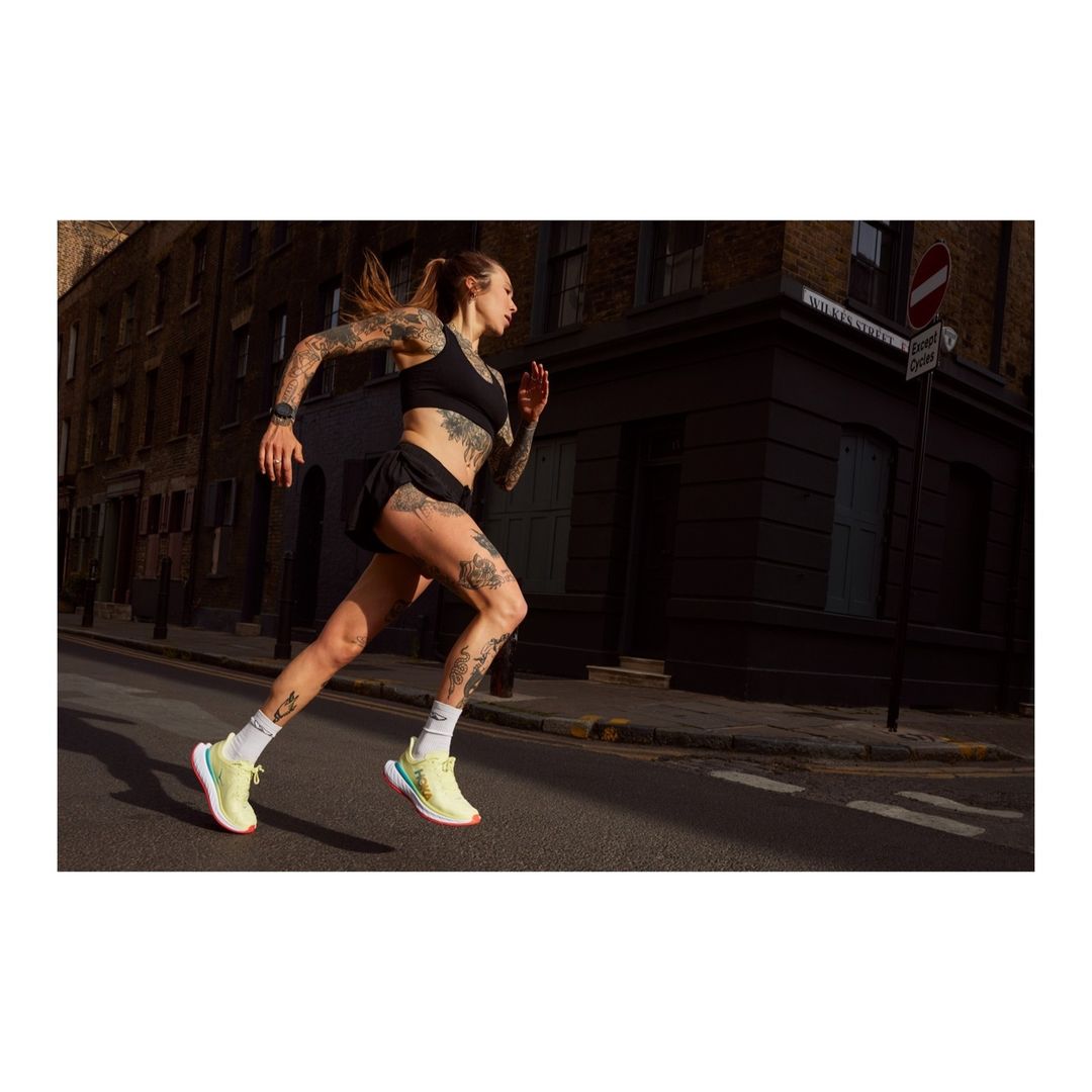 A tattooed woman in sportswear runs on a city street with buildings in the background, showcasing fitness and urban athleticism.