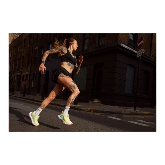 A tattooed woman in sportswear runs on a city street with buildings in the background, showcasing fitness and urban athleticism.