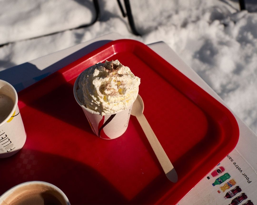 Hot chocolate with whipped cream and cinnamon on a red tray, served outdoors with a snowy background, presented in a bright, natural light.