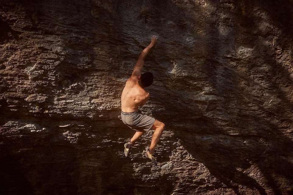 A man climbs a rugged rock face in the outdoors, showcasing strength and determination amidst textured rock and dappled sunlight.