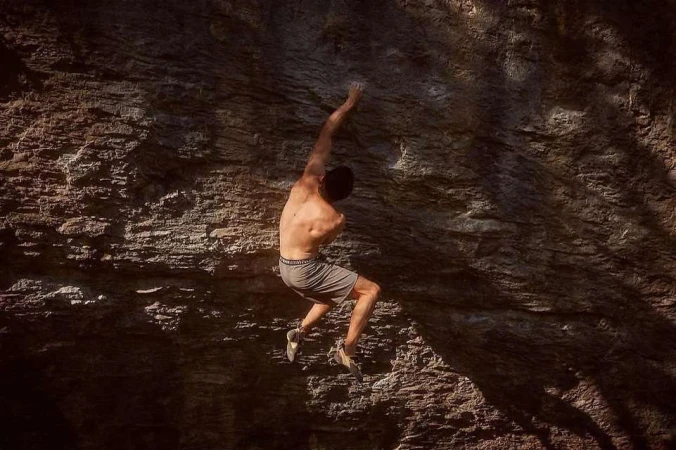 A man climbs a rugged rock face in the outdoors, showcasing strength and determination amidst textured rock and dappled sunlight.