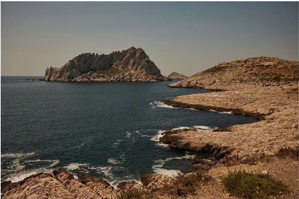 Scenic view of a rocky island, blue water, and coastline with tan-colored rocks and sparse vegetation, set under a clear sky.