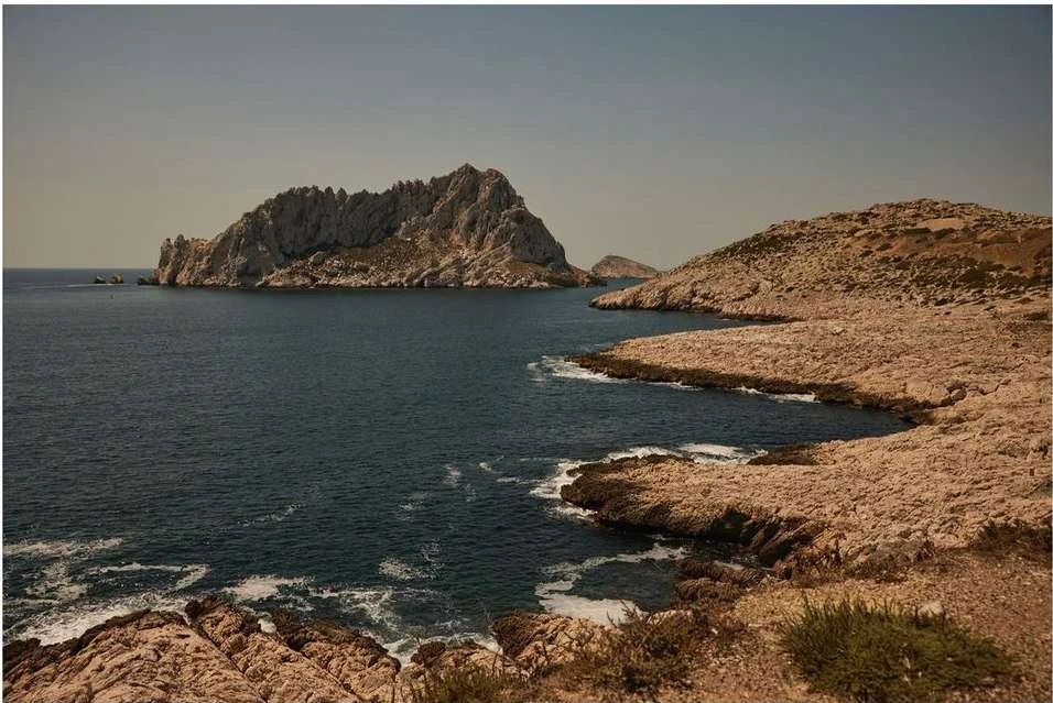 Scenic view of a rocky island, blue water, and coastline with tan-colored rocks and sparse vegetation, set under a clear sky.