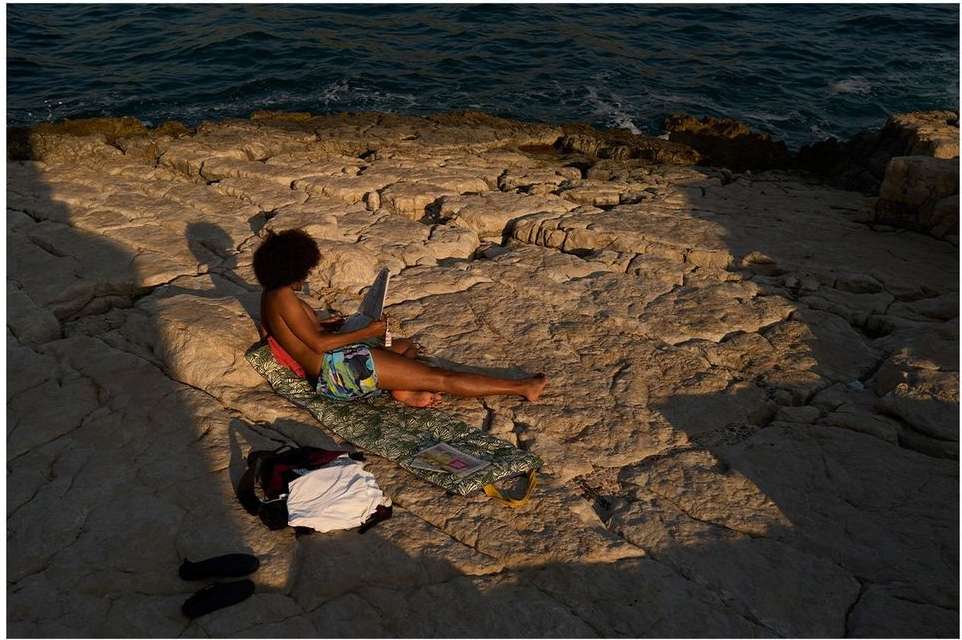 Adult sits on rocky shore with laptop, bag, and towel against a backdrop of ocean water under a warm, sunny sky. Outdoor office concept.