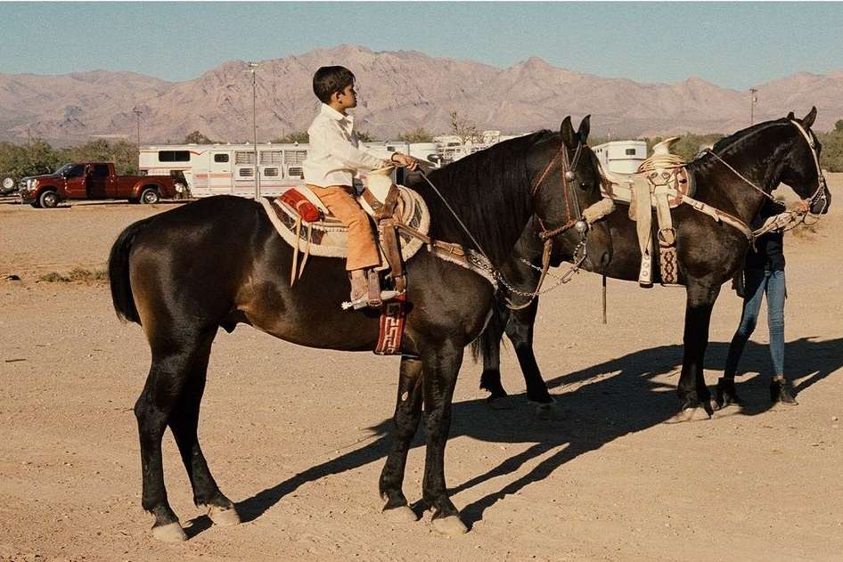 Young boy on a saddled dark brown horse with mountains and an RV in the background.  A second horse is visible to the right.