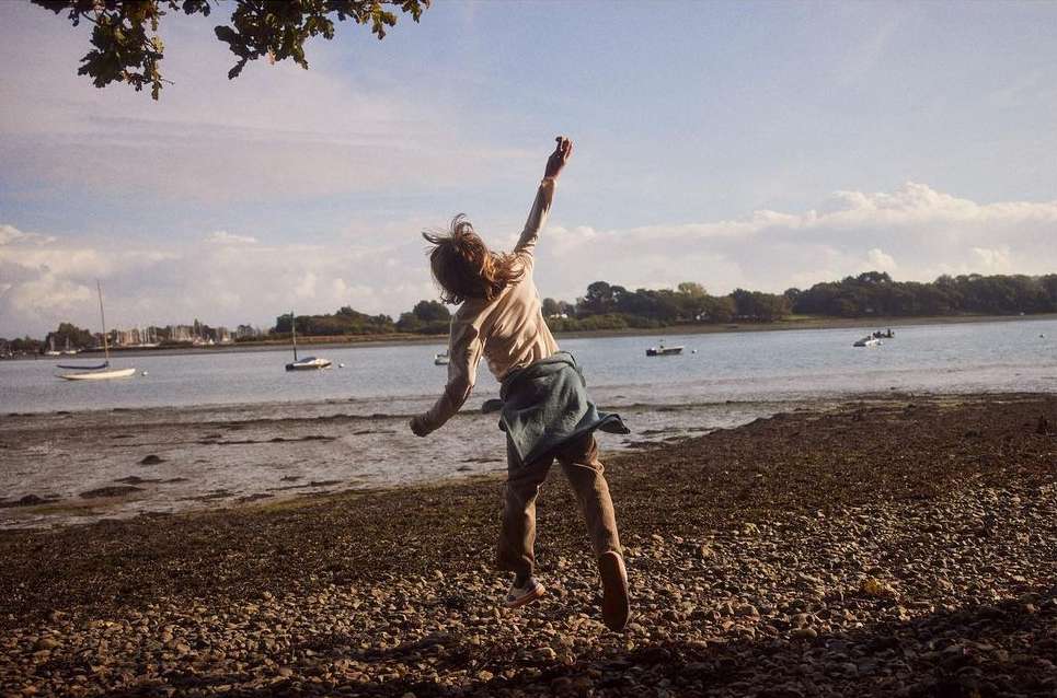 A child jumps on a rocky beach with arms raised, with the ocean and sailboats in the background on a clear day.