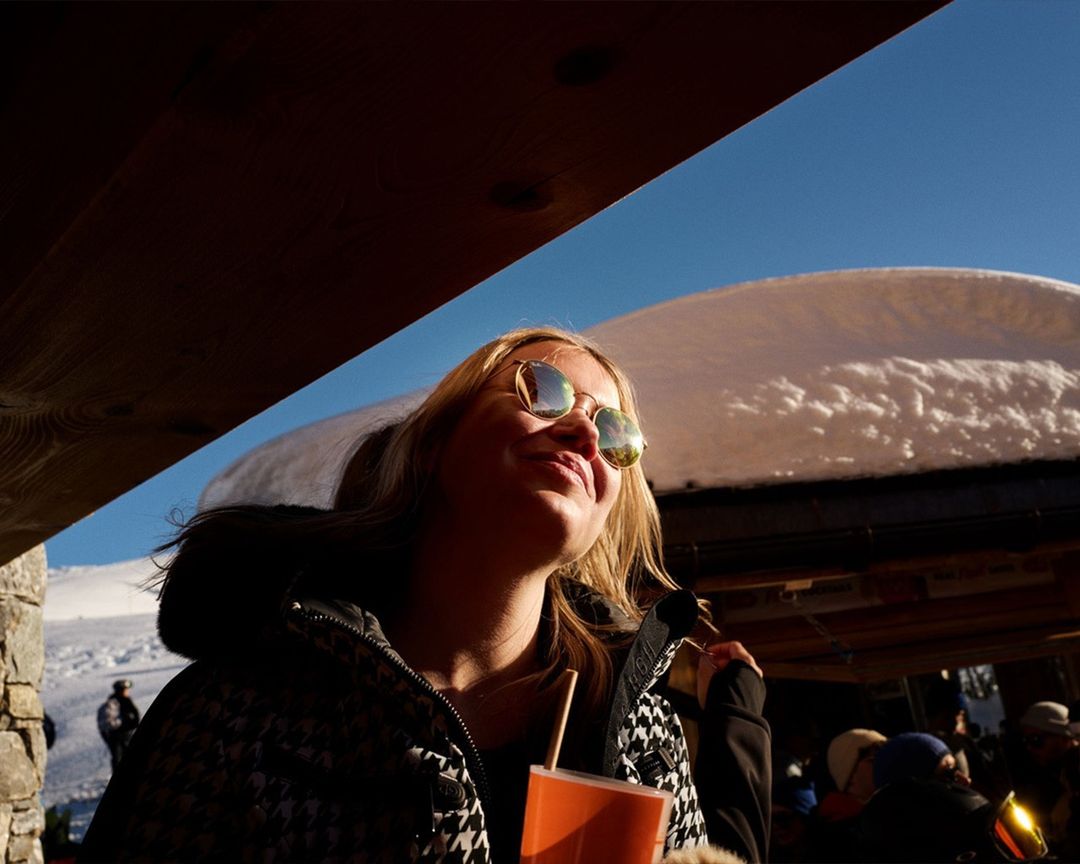 Woman with sunglasses drinks from an orange cup outdoors in winter, with snowy mountains and a blue sky partially visible in the background.