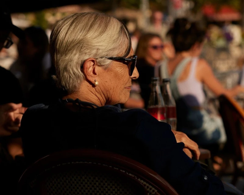 Elegant senior woman with short grey hair and sunglasses sitting at an outdoor cafe, enjoying a relaxed moment in warm sunlight.