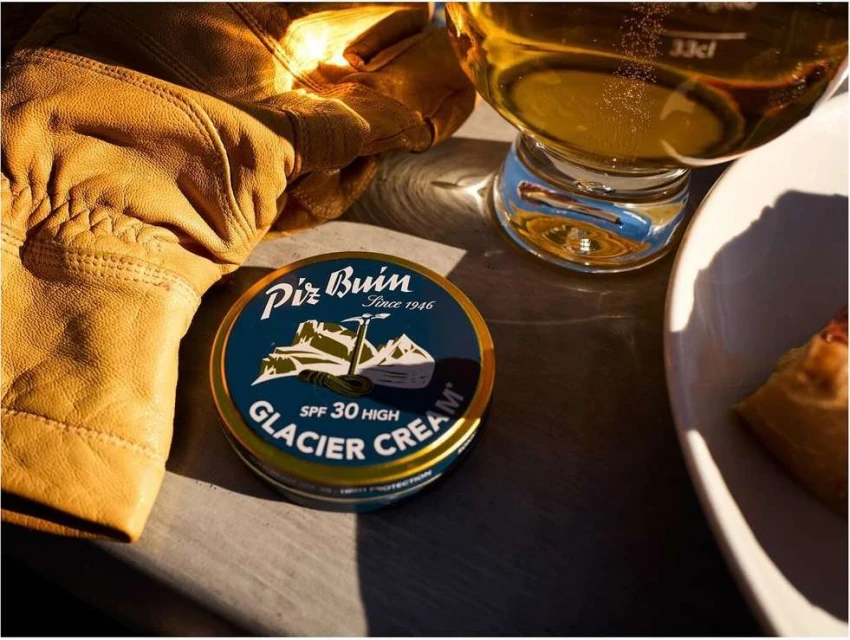 Close-up of Piz Buin Glacier Cream, set near a beer glass and leather gloves on a wooden surface, capturing a moment of outdoor skincare.