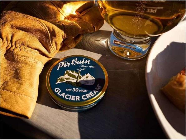 Close-up of Piz Buin Glacier Cream, set near a beer glass and leather gloves on a wooden surface, capturing a moment of outdoor skincare.