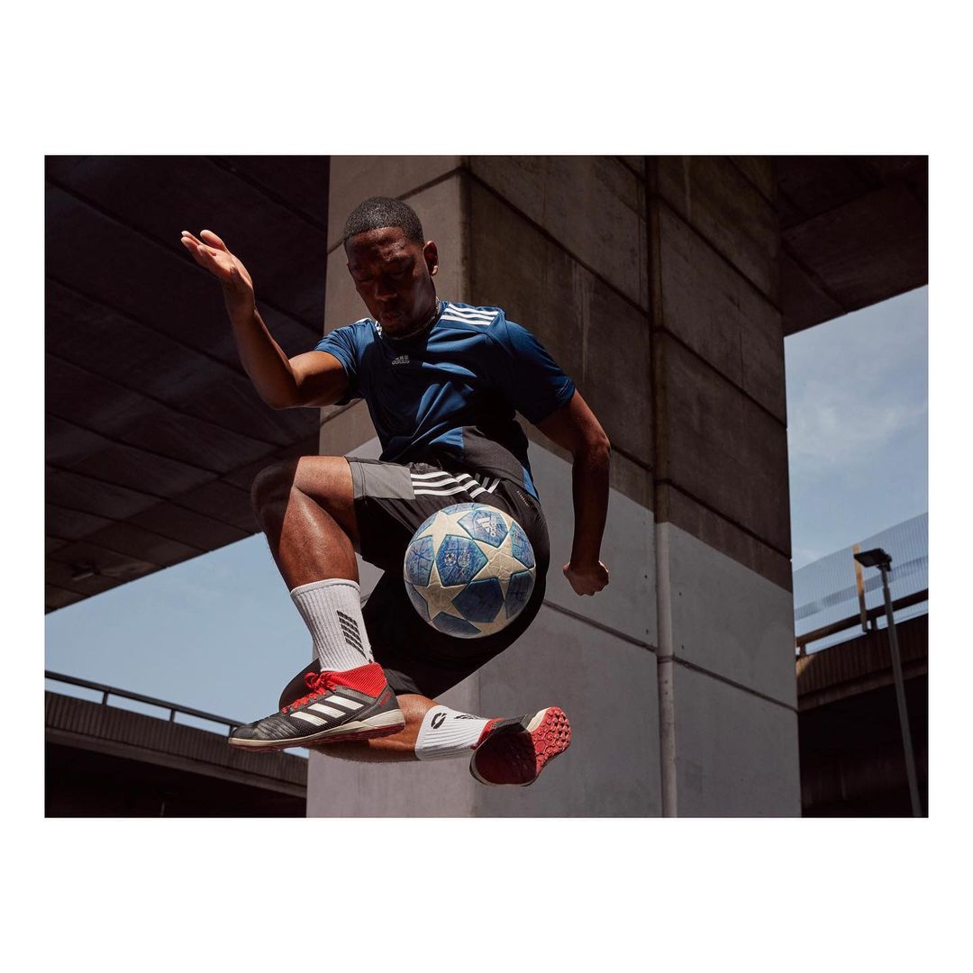 A man in athletic wear jumps in the air kicking a soccer ball with his knee in an urban setting with bright natural lighting.