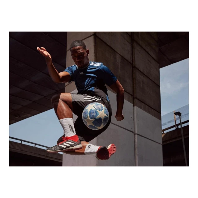 A man in athletic wear jumps in the air kicking a soccer ball with his knee in an urban setting with bright natural lighting.
