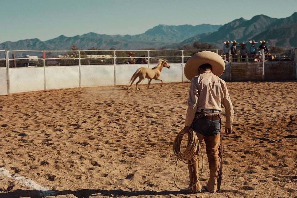 Man in charro outfit holding rope looks at horse inside arena at rodeo against mountain backdrop on a sunny day. Outdoor scene.