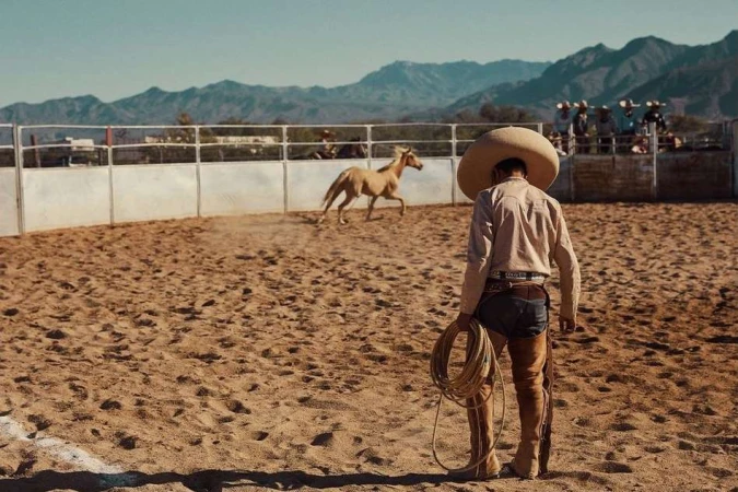 Man in charro outfit holding rope looks at horse inside arena at rodeo against mountain backdrop on a sunny day. Outdoor scene.