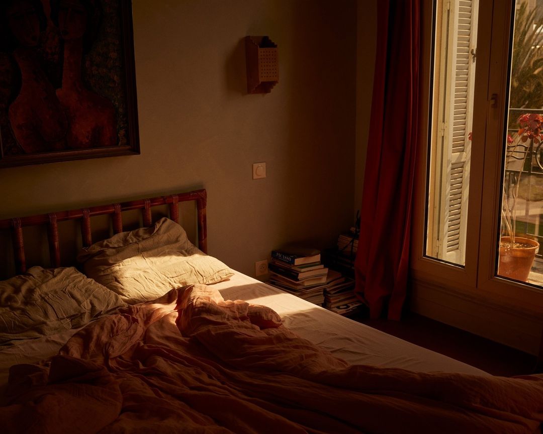 Sunlit bedroom interior with a made bed, artwork on the wall, and a view through an open window to foliage outside.