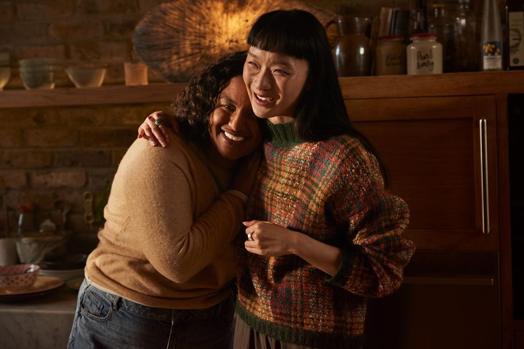 Two smiling women embracing indoors, one with dark curly hair in a tan sweater, the other with straight black hair in a plaid sweater.