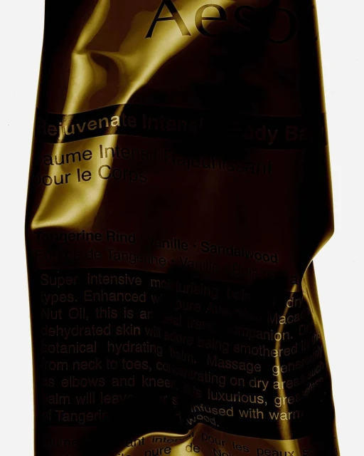 Close-up shot of an Aesop Rejuvenate Intensive Body Balm tube with a gold metallic finish, showcasing the product details and branding.