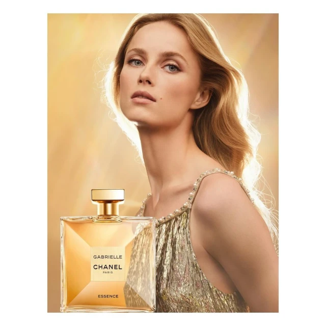 Close-up shot of a blonde adult woman and a bottle of Gabrielle Chanel Essence perfume in a studio with gold background lighting.