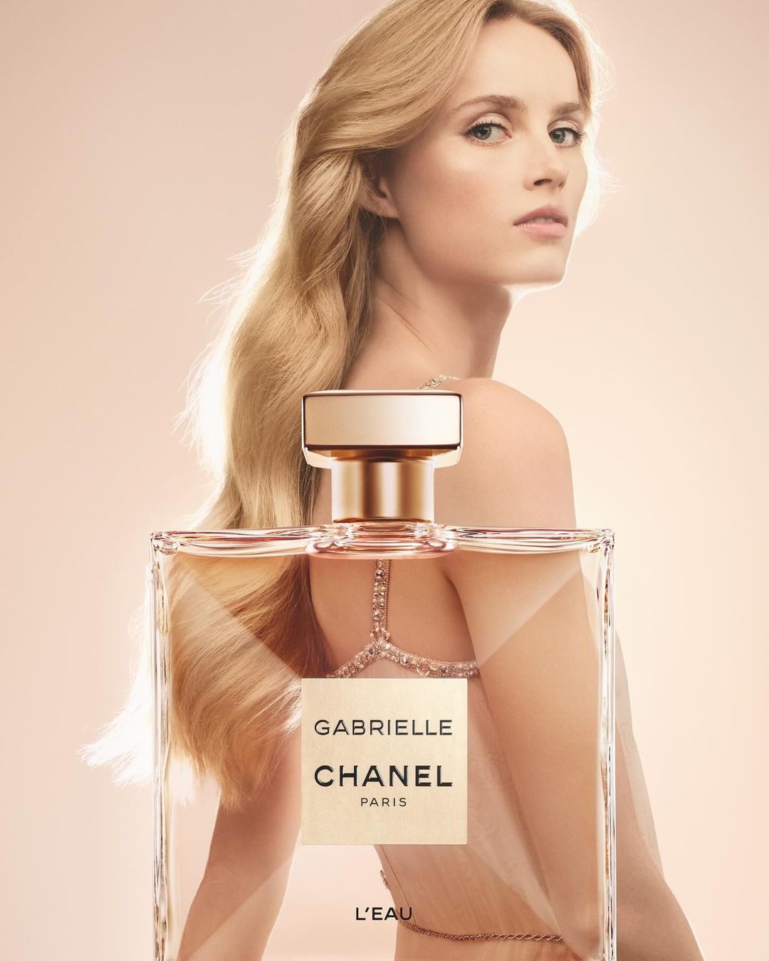 Blonde model with Chanel Gabrielle L'Eau perfume, soft peach backdrop, shoulder pose, long flowing hair, and subtle jewelry.