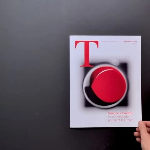 Magazine cover featuring red and black graphic design on a white background, held in a hand against a dark gray backdrop.