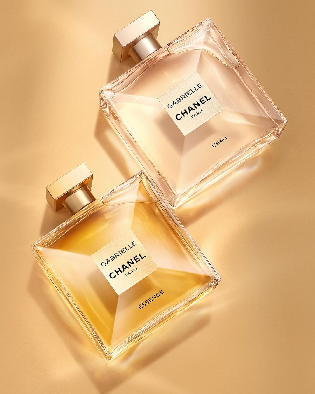 Two Gabrielle Chanel perfume bottles, one 'Essence' and one 'L'Eau,' with square bottles and gold accents, on a warm-toned background.