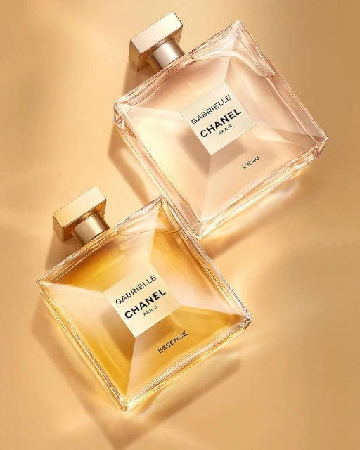 Two Gabrielle Chanel perfume bottles, one 'Essence' and one 'L'Eau,' with square bottles and gold accents, on a warm-toned background.