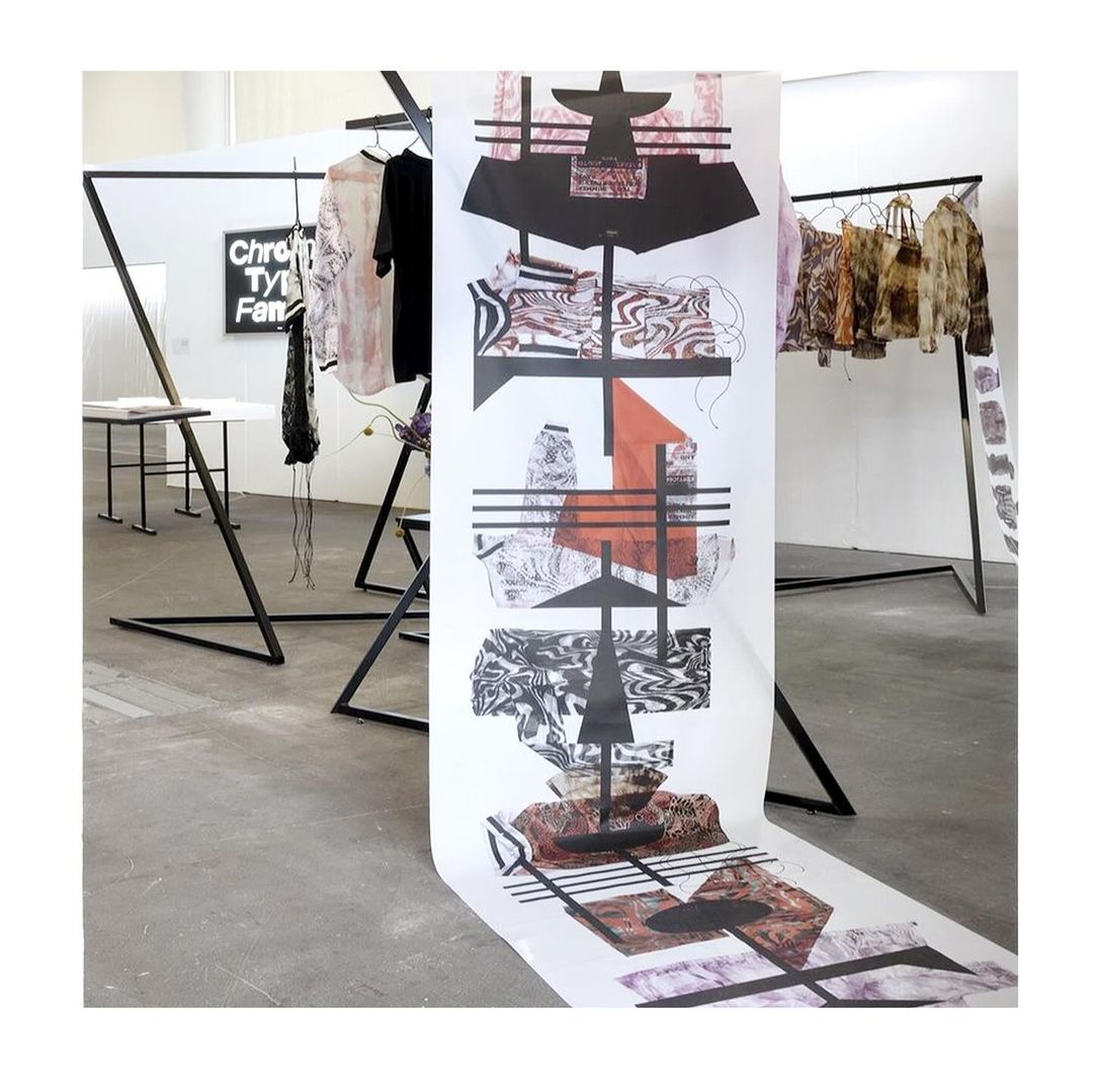 Art installation featuring black metal clothing rack with garments and a large printed art piece in a white, modern gallery space.