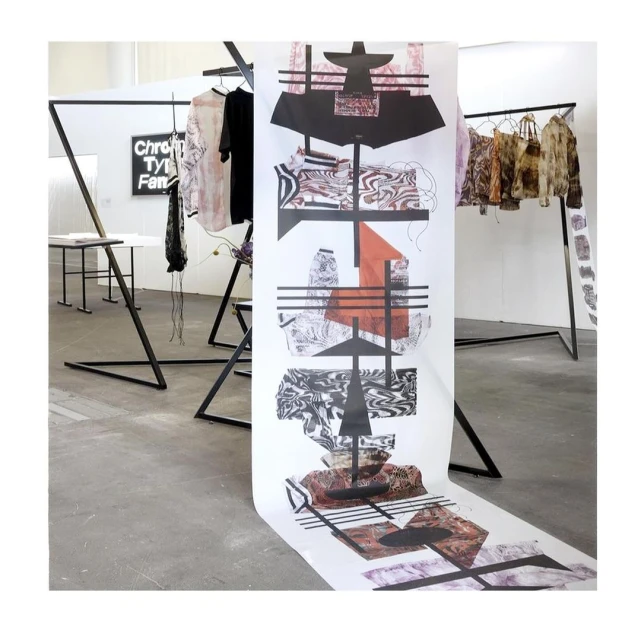 Art installation featuring black metal clothing rack with garments and a large printed art piece in a white, modern gallery space.