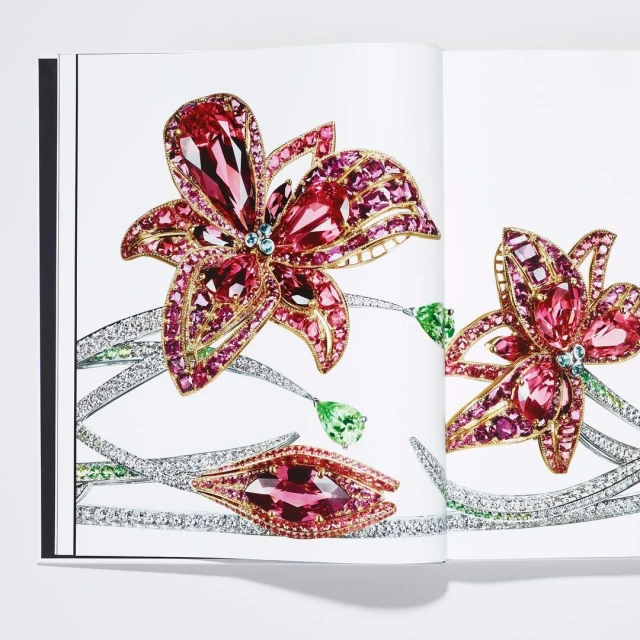 Elegant floral jewelry designs featured in open book. Pink and gold flower brooches, diamond and green gemstone accents on white.