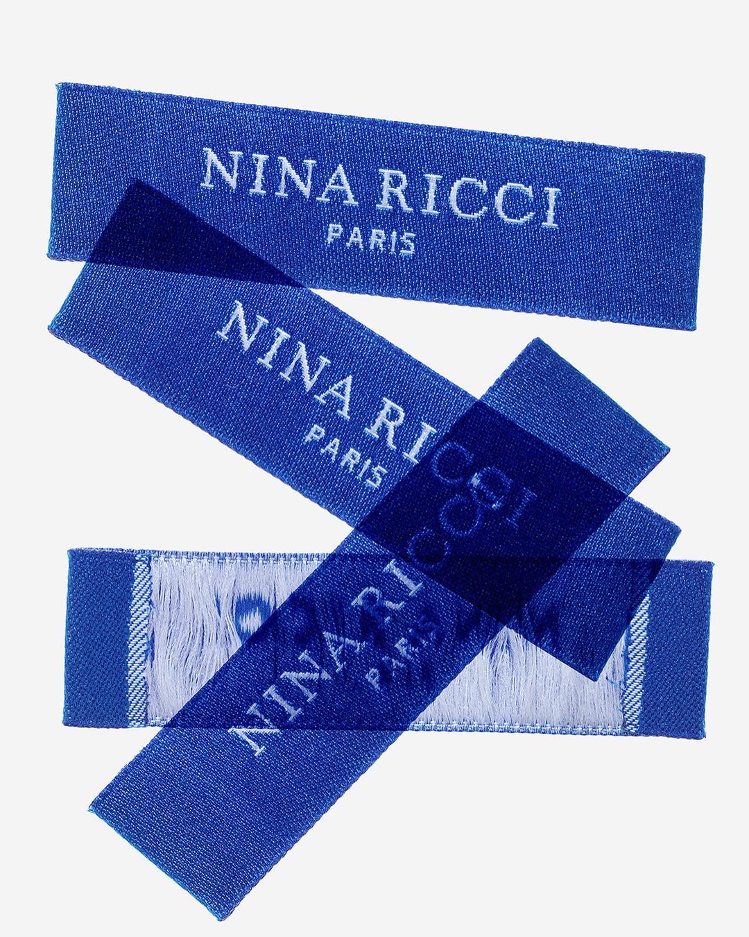 Three blue fabric labels with the 'Nina Ricci Paris' brand name stacked diagonally against a bright white background in a studio shot.