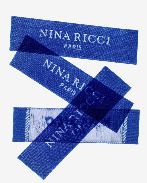 Three blue fabric labels with the 'Nina Ricci Paris' brand name stacked diagonally against a bright white background in a studio shot.