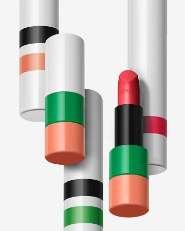 Hermès lipstick tubes in various colors and patterns, one with the coral lipstick extended, studio shot on a clean white background.