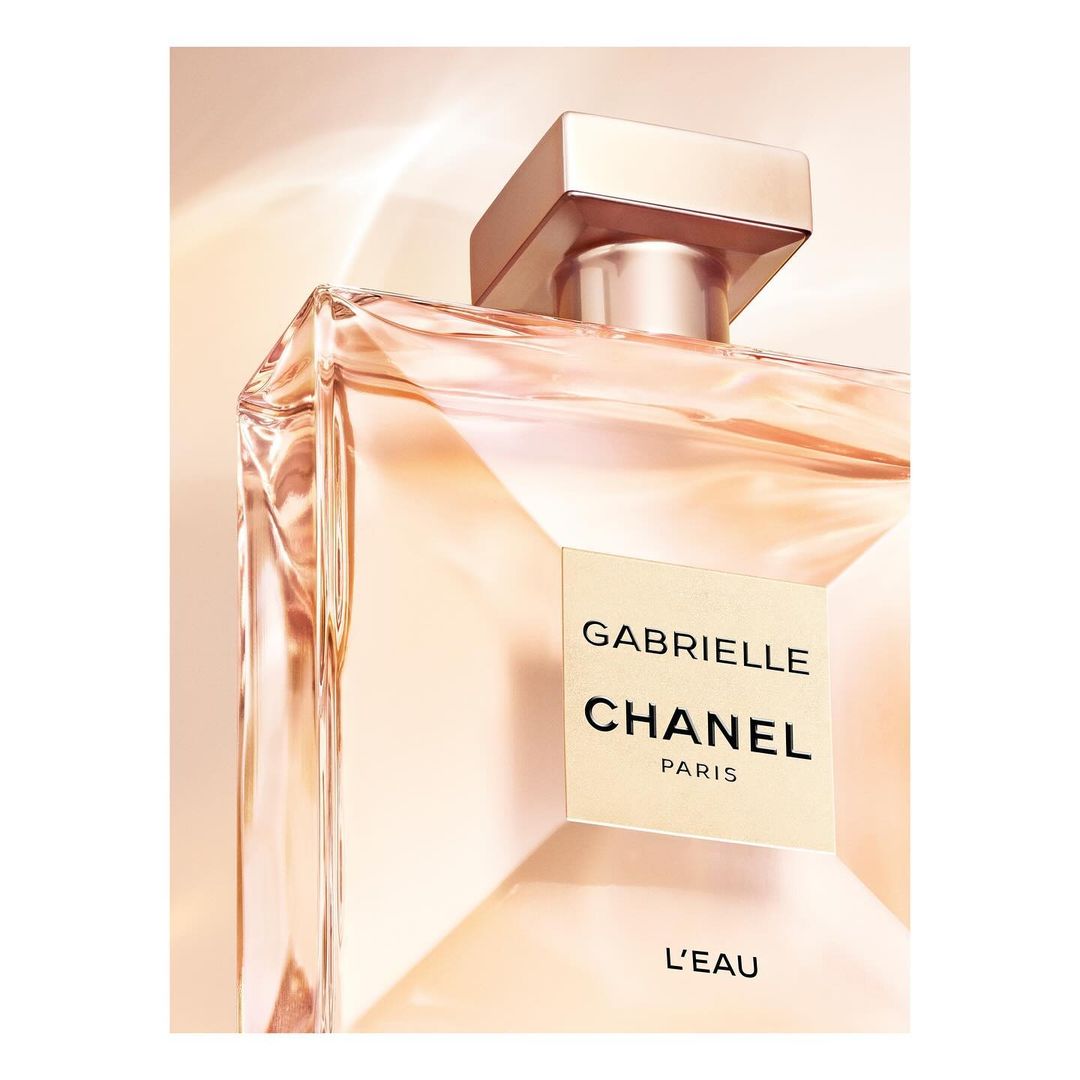 Close-up of a light pink-tinted glass perfume bottle, featuring a square gold cap and a label reading 'Gabrielle Chanel Paris L'Eau'.