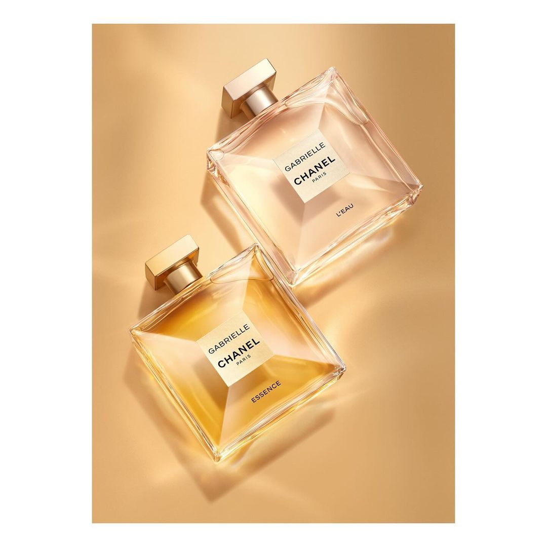Two square Chanel perfume bottles on a gold surface, one containing clear essence and the other containing golden essence.