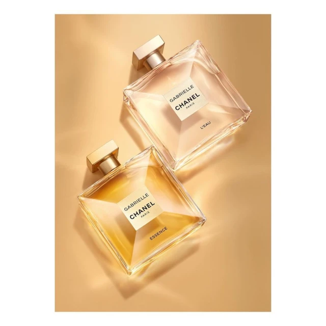 Two square Chanel perfume bottles on a gold surface, one containing clear essence and the other containing golden essence.