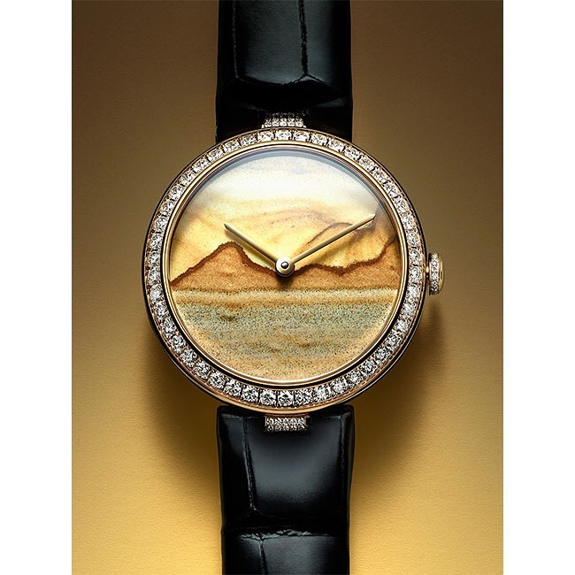 Close-up of a luxury wristwatch featuring a landscape dial with golden mountains and a black leather strap against a gold backdrop.