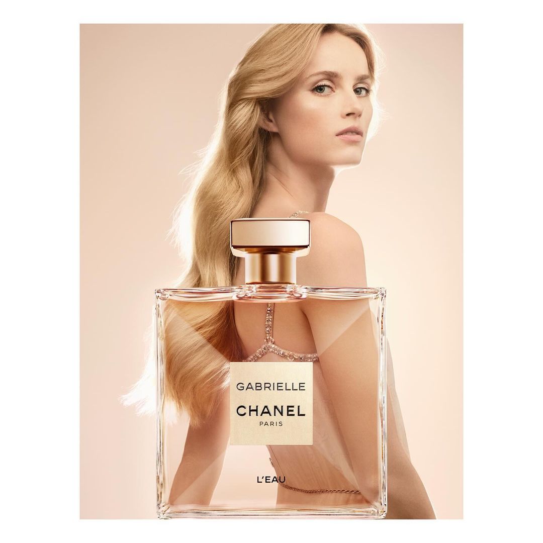 Glamorous woman showcasing Gabrielle Chanel L'Eau perfume. Bottle in the foreground, long blonde hair, sophisticated aesthetic, light background.