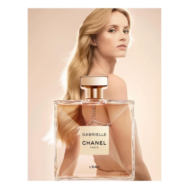 Glamorous woman showcasing Gabrielle Chanel L'Eau perfume. Bottle in the foreground, long blonde hair, sophisticated aesthetic, light background.