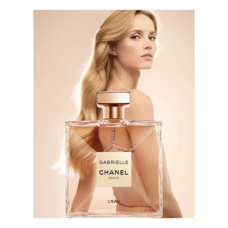 Glamorous woman showcasing Gabrielle Chanel L'Eau perfume. Bottle in the foreground, long blonde hair, sophisticated aesthetic, light background.