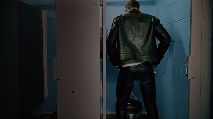 Back view of a man in black leather jacket and pants standing in a public bathroom stall, facing away from the camera.