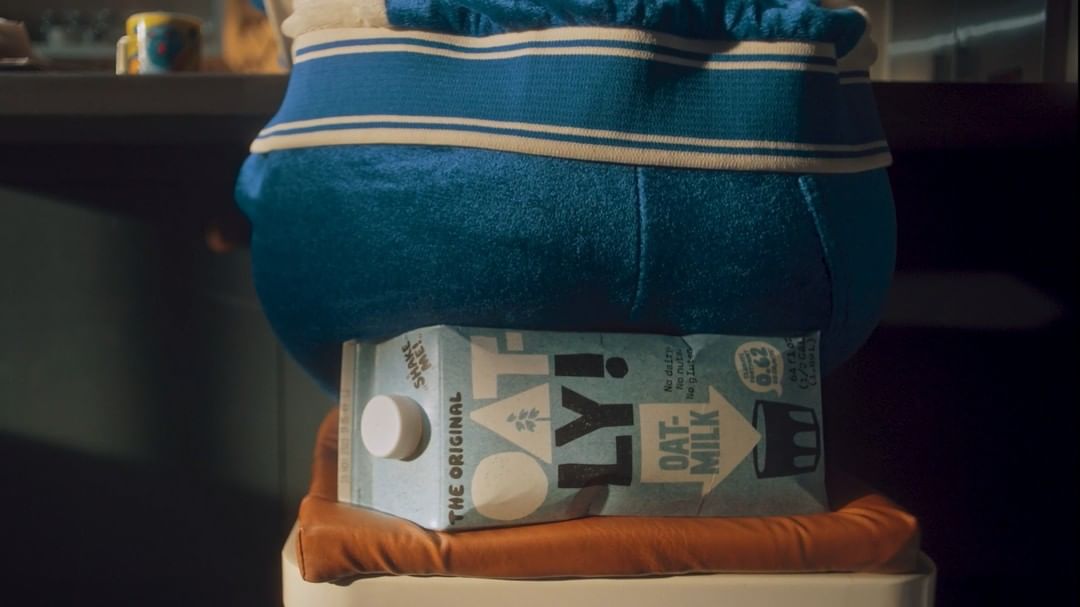 Oatly oat milk carton sits on orange cushion with blue and white striped fabric behind in an indoor setting with dark background.