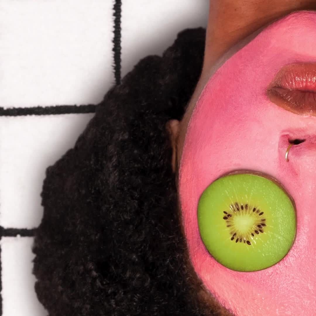 Close-up of a face with a pink face mask and a slice of kiwi over the eye, set against a white tiled background.