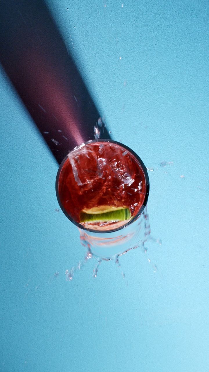 Overhead view of a red cocktail in a glass with ice and lime, liquid splashing against a light blue background with a long shadow.