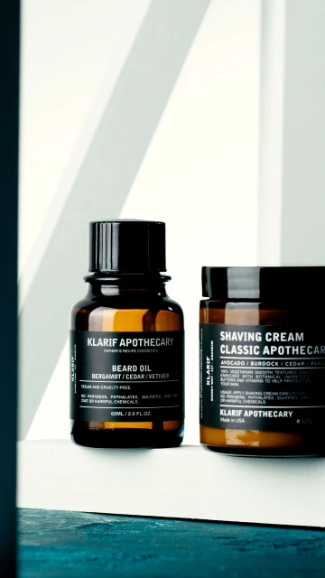 Product shot of Klairf Apothecary beard oil and shaving cream, displayed in glass and plastic containers with black labels.