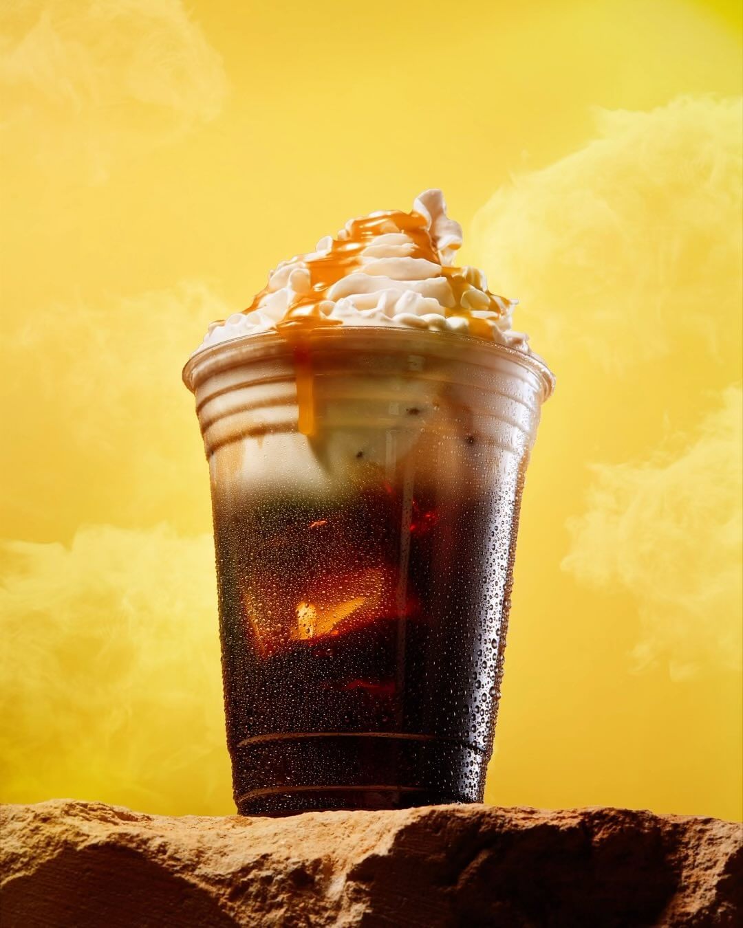 Iced coffee in a plastic cup topped with whipped cream and caramel drizzle sitting on a rock against a bright yellow background.