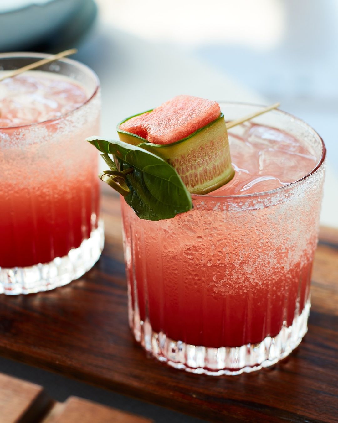 Two refreshing cocktails with ice, garnished with watermelon, cucumber, and a green leaf, served on a wooden tray.